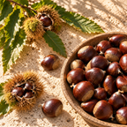 A wooden bowl filled with brown chestnuts and a few spiky brown husks, arranged on sandy ground with green leaves in warm sunlight.